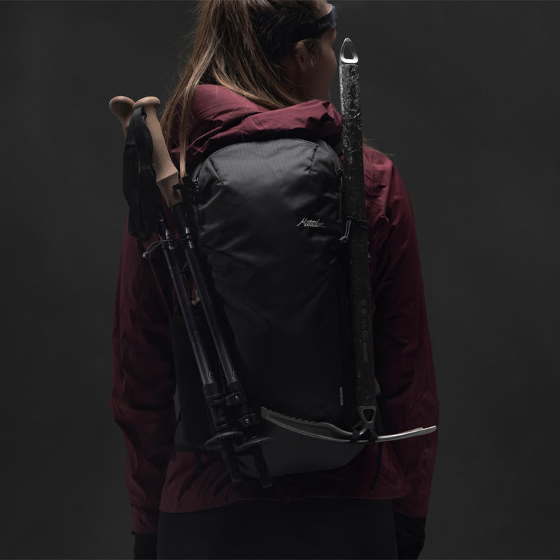 Back view of woman on gray background wearing Beast18 with trekking poles and ice axe attached