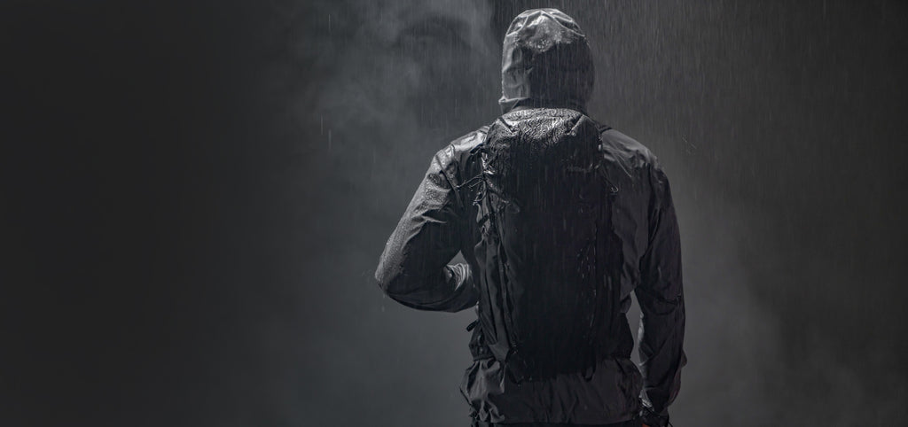 Back view of person in gray raincoat wearing black backpack in black, rainy, moody scene