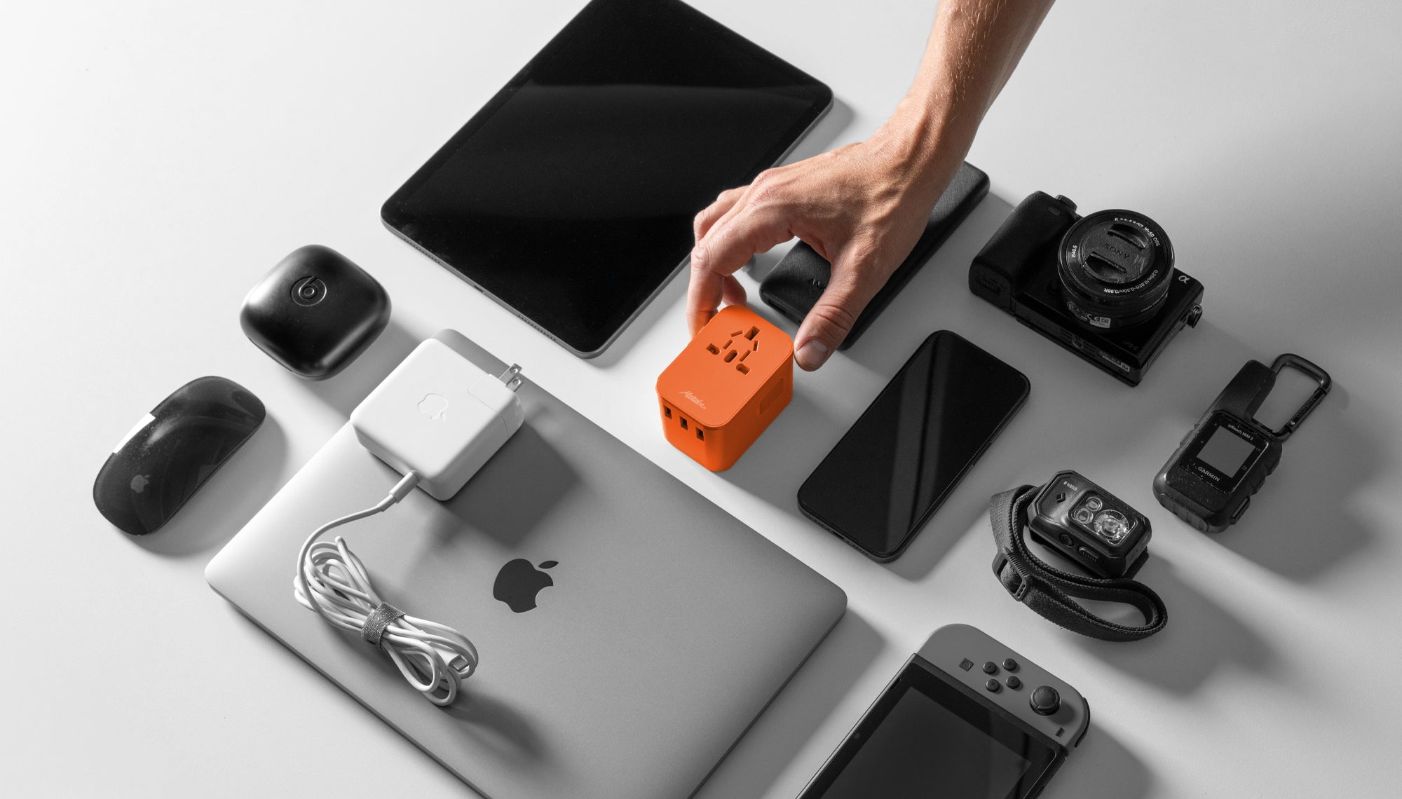 Hand placing orange adapter in group of electronics including laptop, phone, camera, head lamp, earbuds, ipad, and game counsel