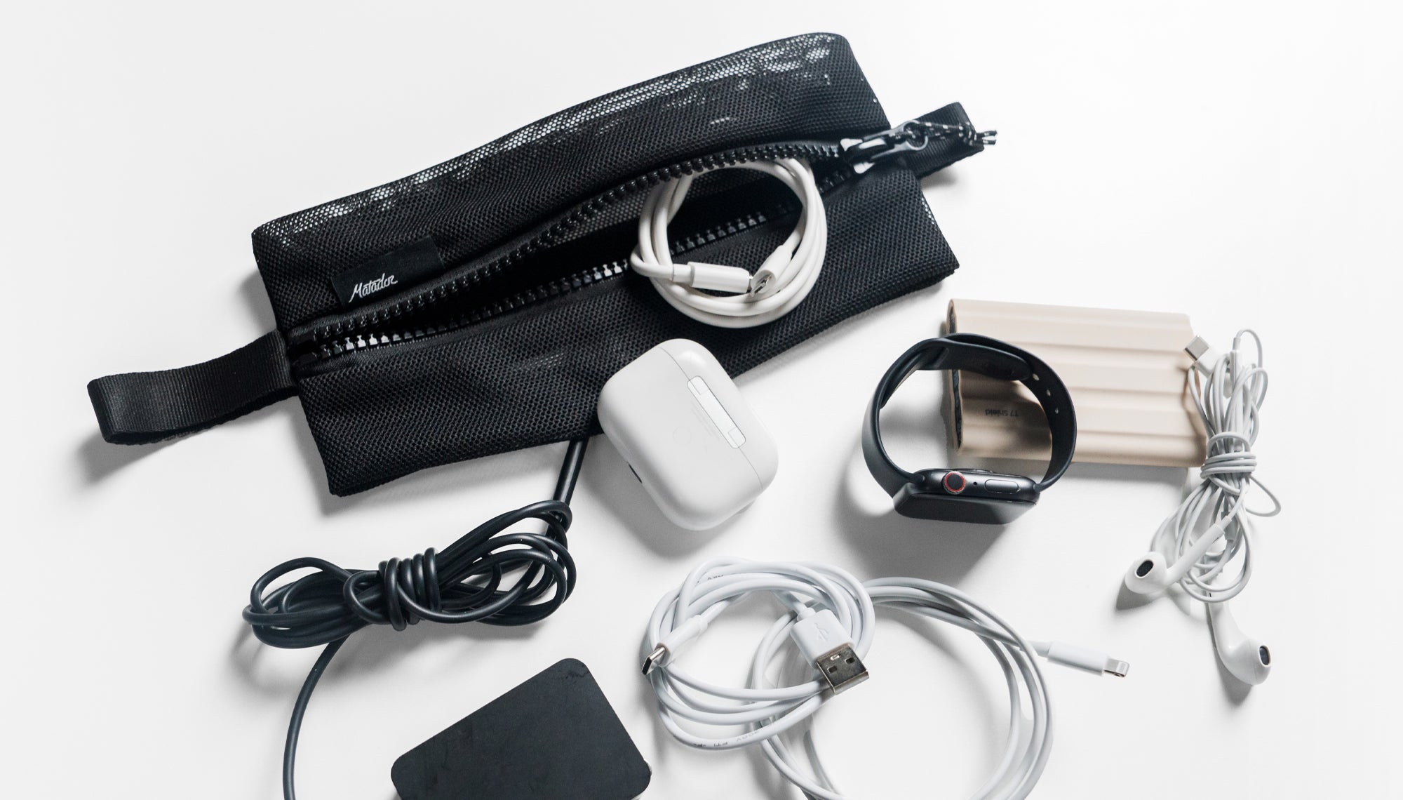 Large mesh bag on white background surrounded by various cords and small electronics like hard drives apple watch, and ear pods