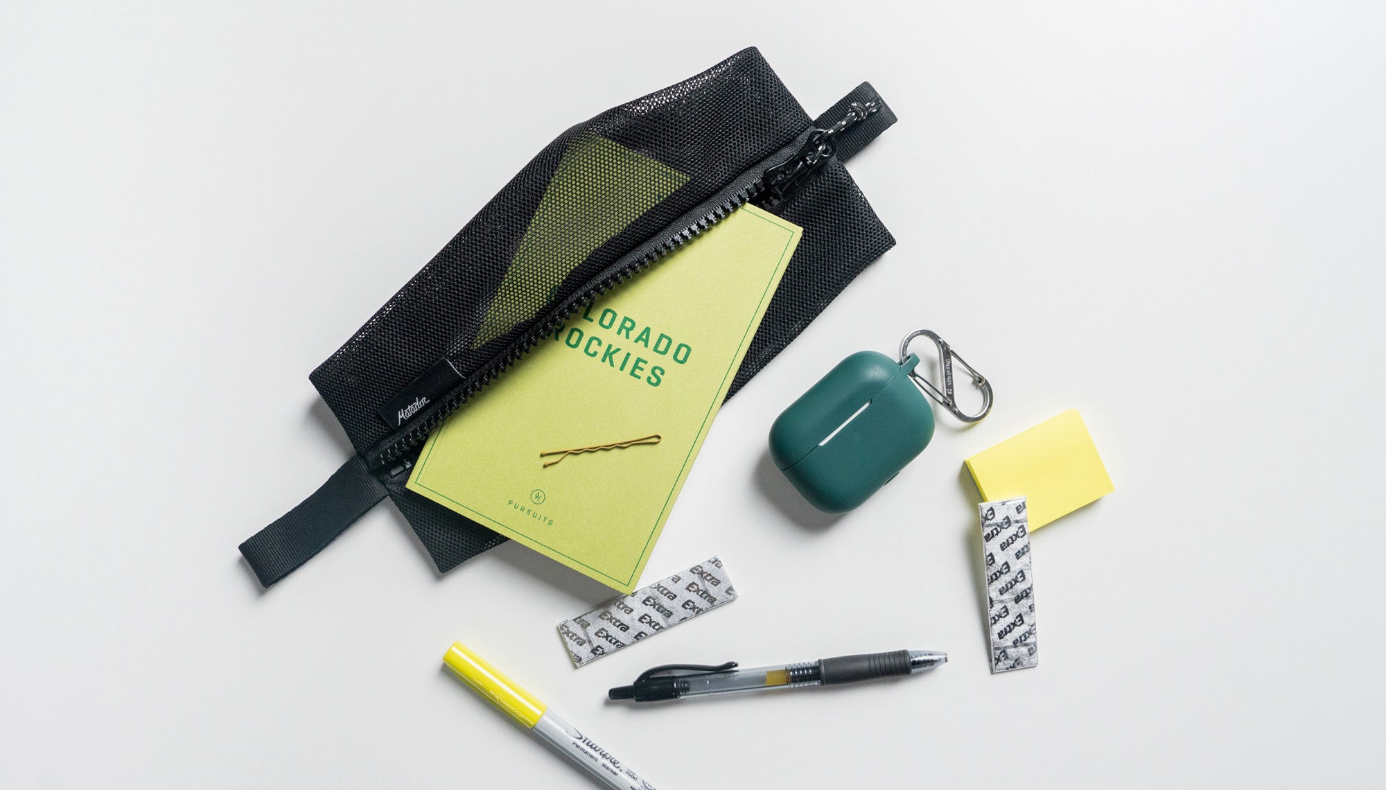 medium mesh bag on white background surrounded by pens, post it notes, gum, earbuds, and a Colorado rockies notebook
