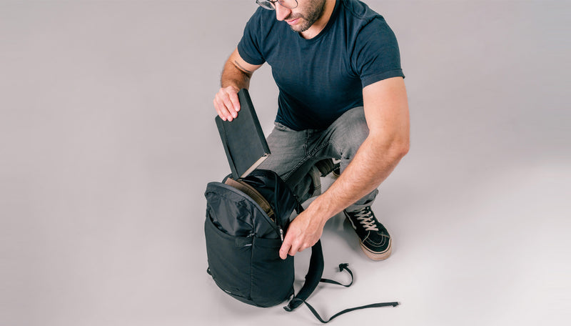 Man kneeling on light gray background placing black notebook into black backpack