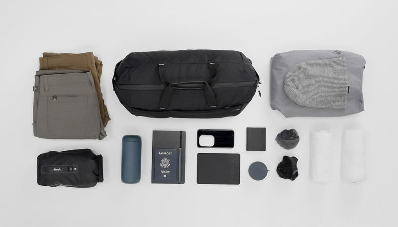 Black duffle on light gray background laid out with various items to go inside: Pants, dry bag, water bottle, note book, passport, phone, headphones, wallet, canister, socks, tees, rain coat, and knit hat