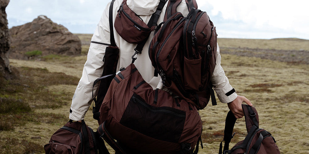 man standing in a grassy field wearing multiple bags and backpacks