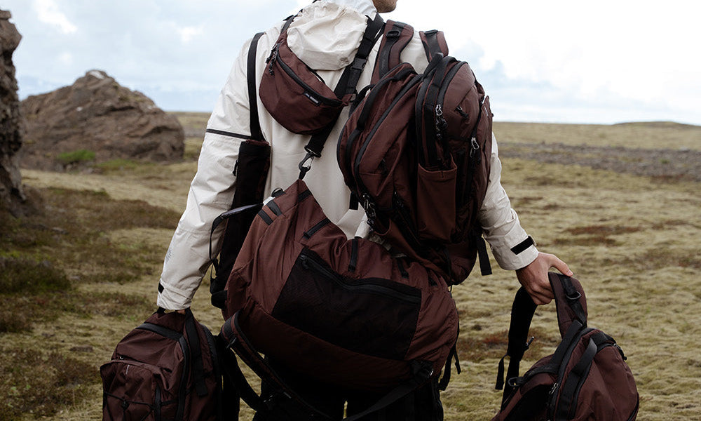 man standing in a grassy field wearing multiple bags and backpacks