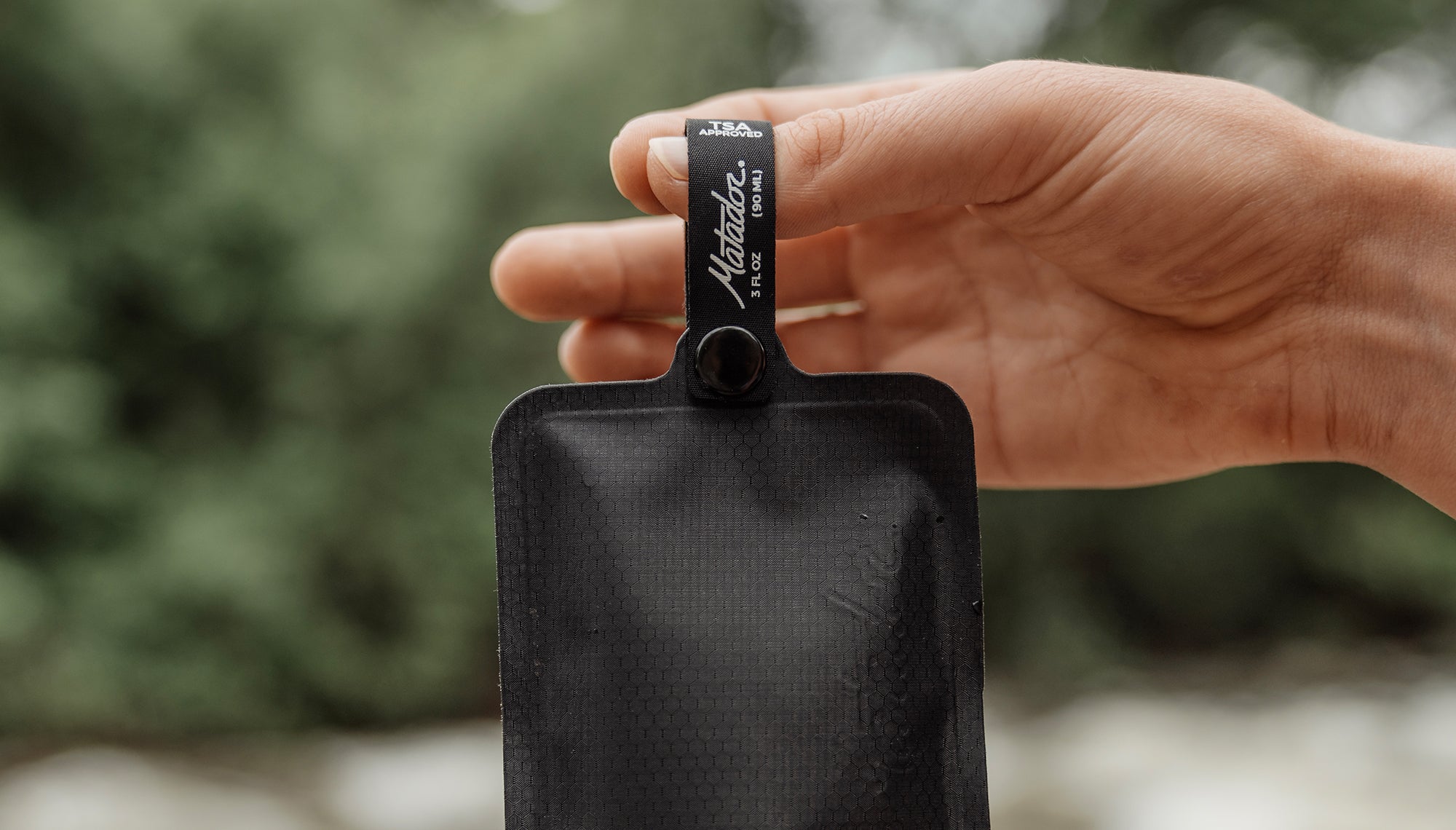 Hand holding FlatPak Toiletry Bottle from loop in front of blurred leafy background