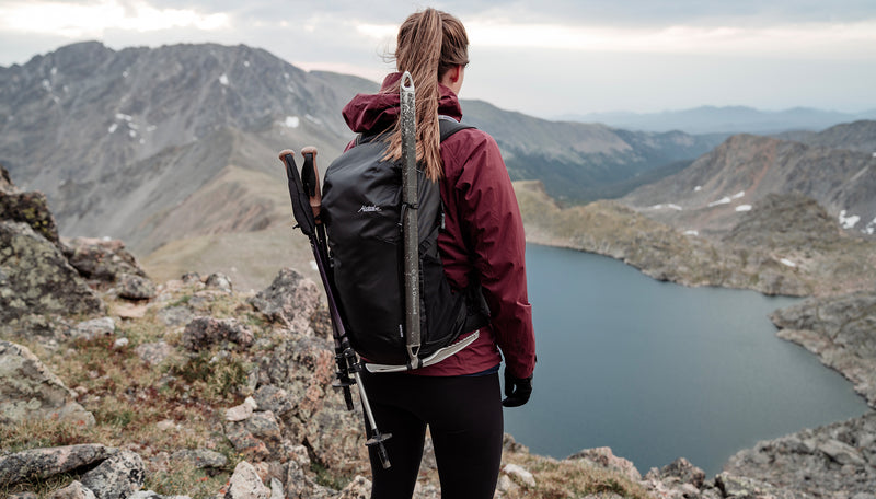 Woman in maroon jacket, wearing Beast18, standing on mountain peak, looking down at alpine lake