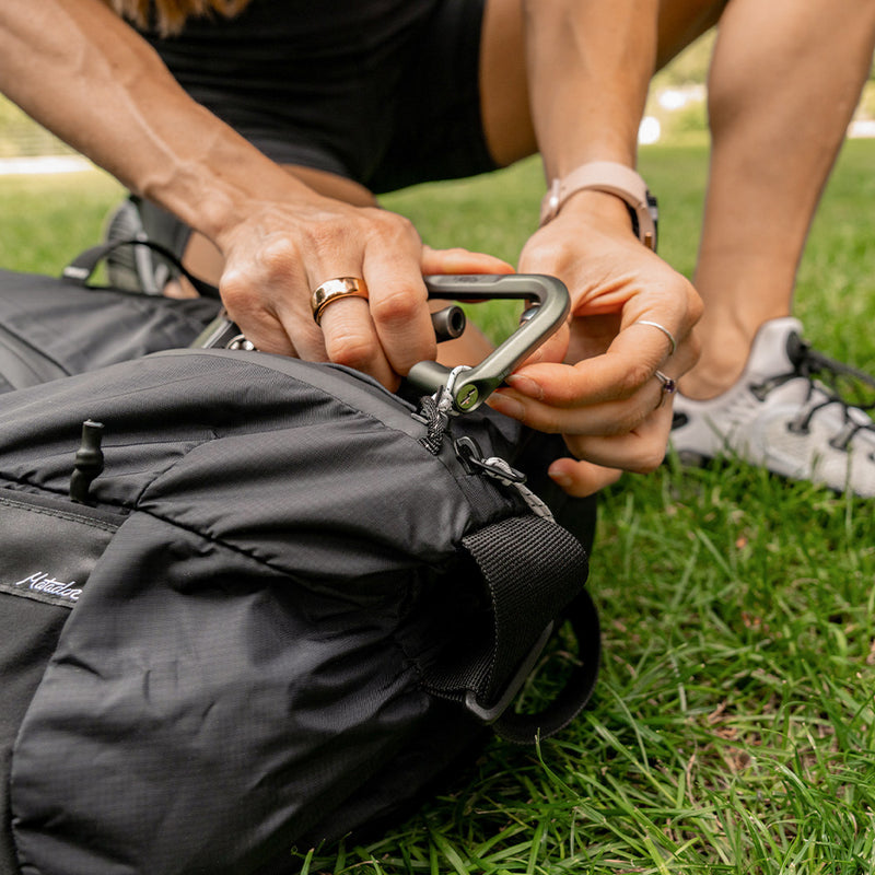 green carabiner being clipped onto black duffle bag sitting on green grass
