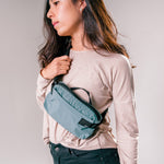 woman on light gray background wearing slate blue sling bag across the front of her body
