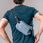 man on light gray background wearing slate blue sling bag across the back of his body