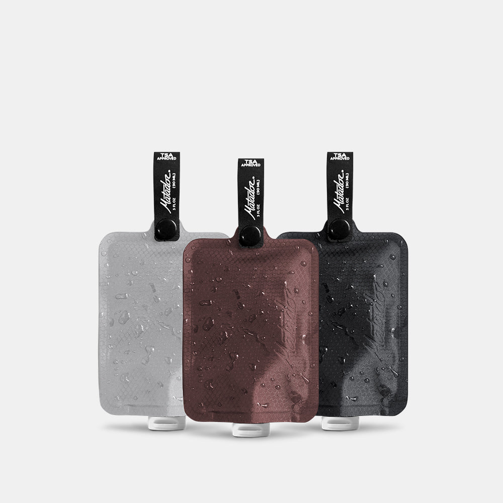 3 multi colored FlatPak Toiletry Bottles—charcoal, garnet, and arctic white—on light gray background