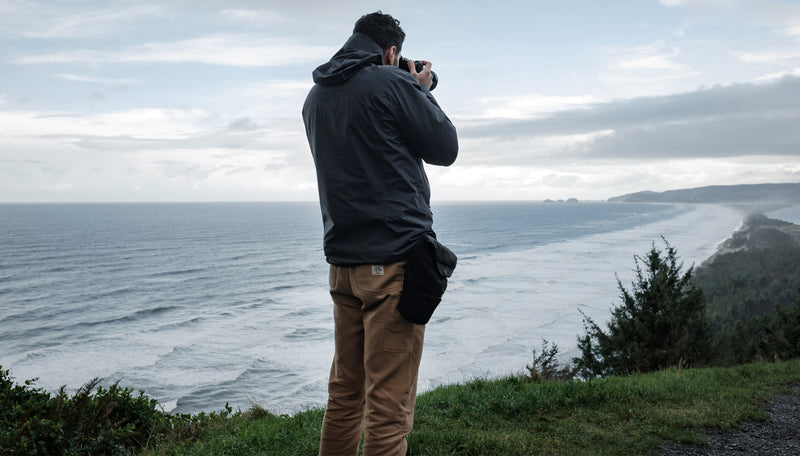 Man on ocean coast, taking a photo while Camera Base Layer is attached to his waistbelt