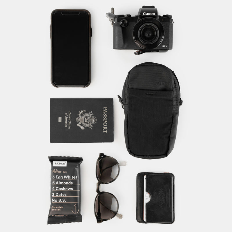 Various items laid out with speed stash: camera, cell phone, passport, RX bar, sunglasses, and wallet