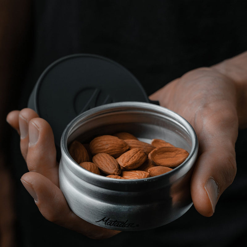 Hand holding open 100ml canister filled with almonds