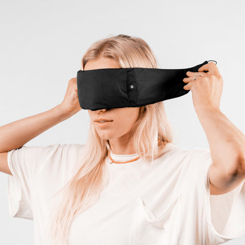 Woman on light gray background, placing black eyemask over her eyes