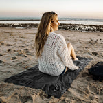 woman on sandy beach, sitting on charcoal beach towel as she views the sunset