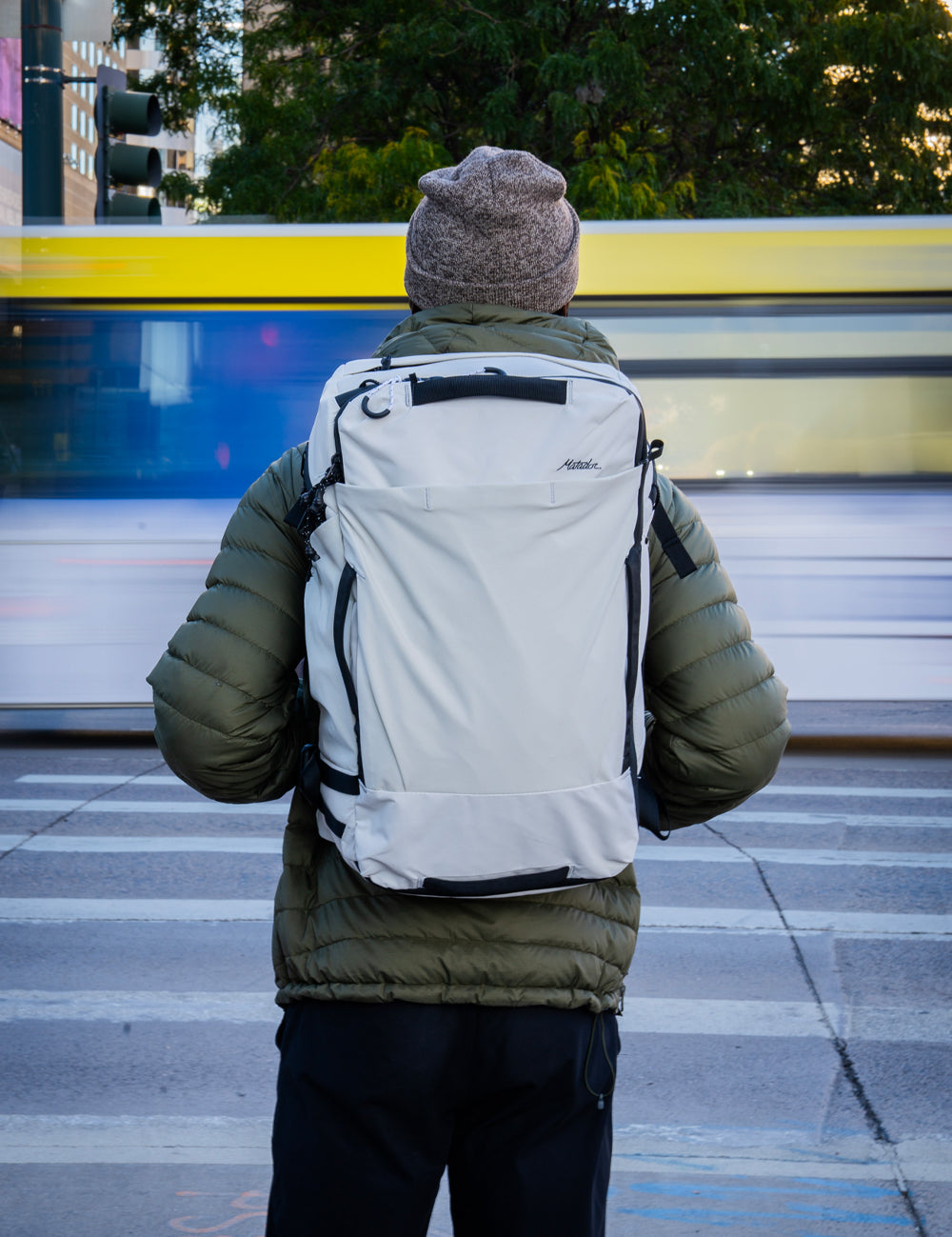 person in green jacket with white backpack standing in front of crosswalk with moving traffic