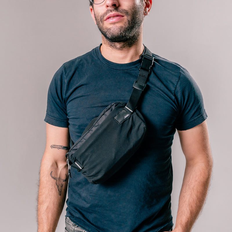 man on light gray background wearing black sling bag across the front of his body