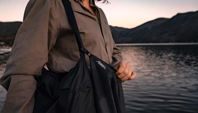 Woman standing at sunset lake, holding NanoDry Towel that hangs off her bag