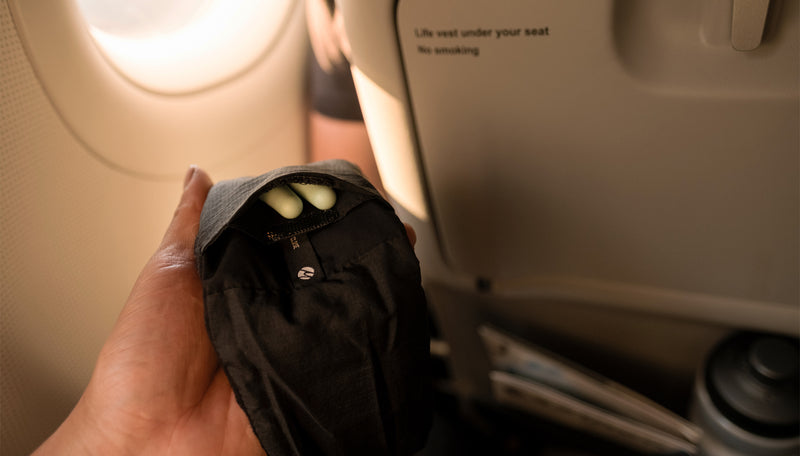 Hand holding sleep mask on airplane with earplugs peeking out from pocket