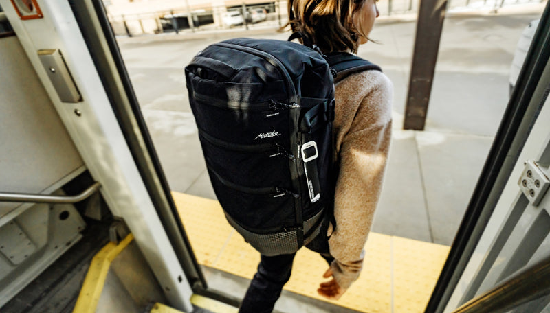 Woman exiting train, wearing black backpack with gear tag hanging on outside.