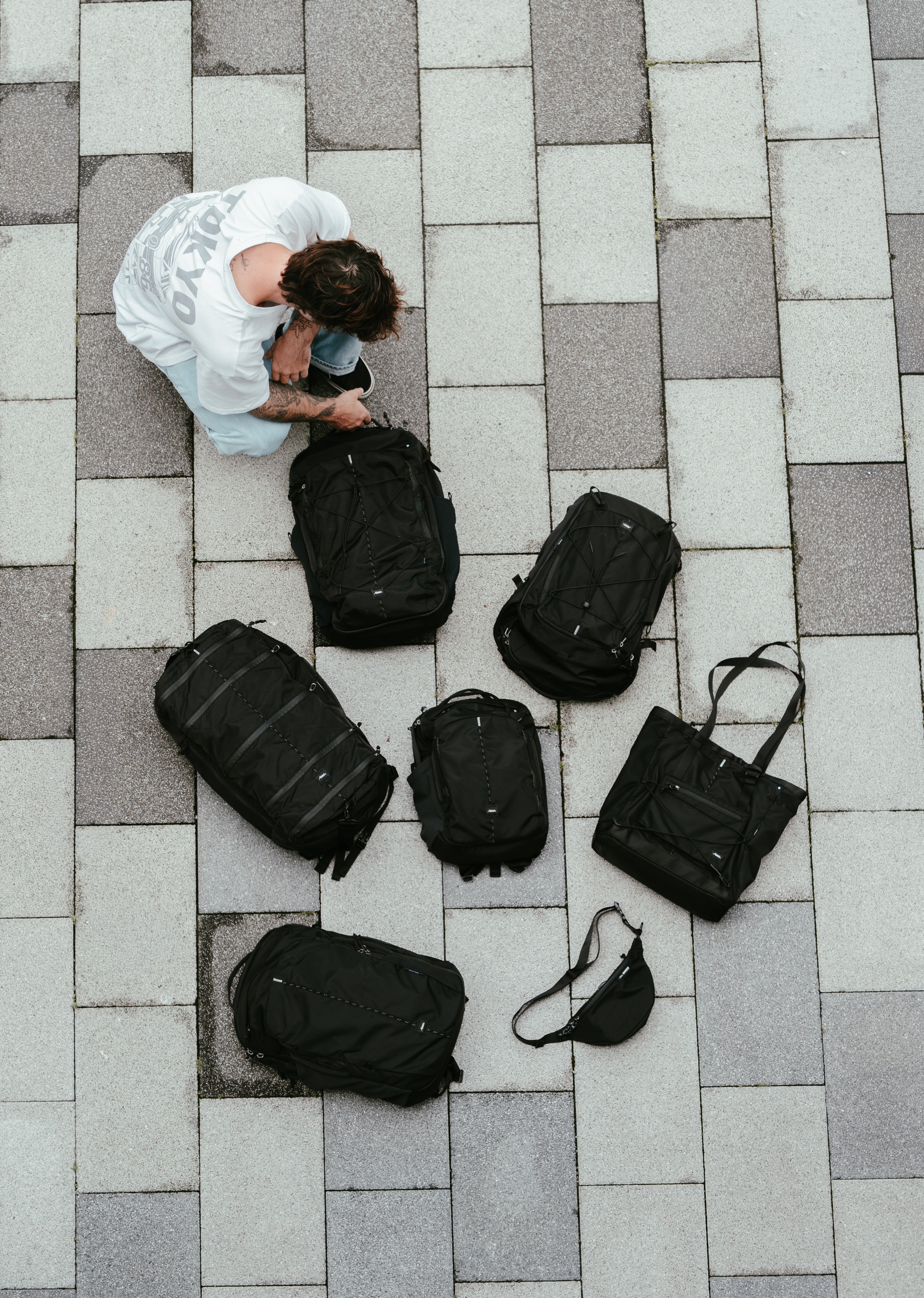 A top-down view of a person crouching beside a group of black backpacks, bags, and a small sling bag out on gray paving stones.