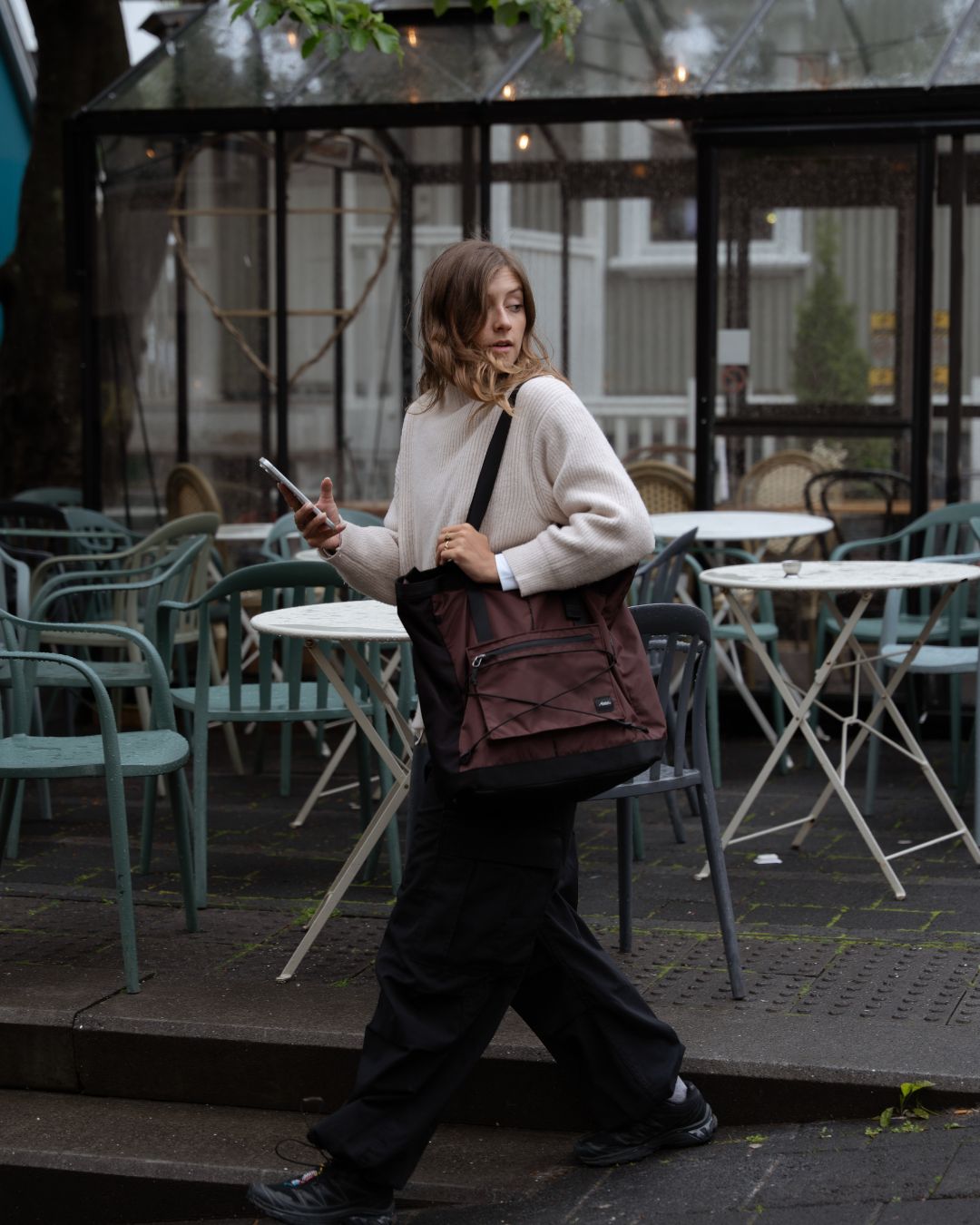 A woman walking outdoors carrying a dark burgundy tote bag on her shoulder, wearing a cream sweater and black pants, with cafe tables and chairs in the background.