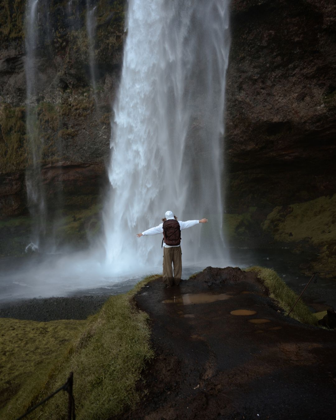A person standing in front of a tall waterfall with arms outstretched, wearing a dark backpack, set against a mossy rock wall.