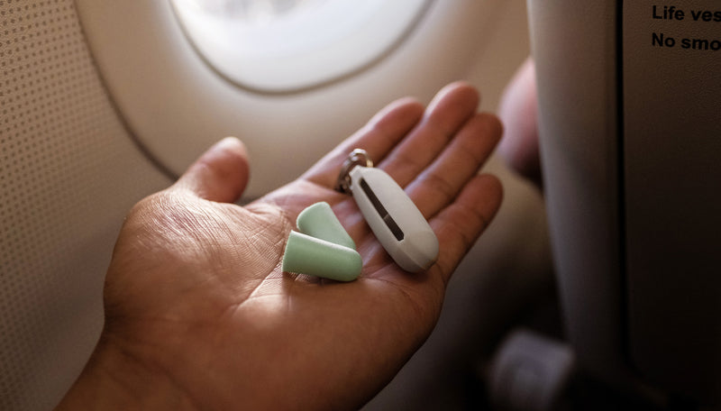 Hands holding white earplugs case and 2 mint colored ear plugs, in front of airplane window