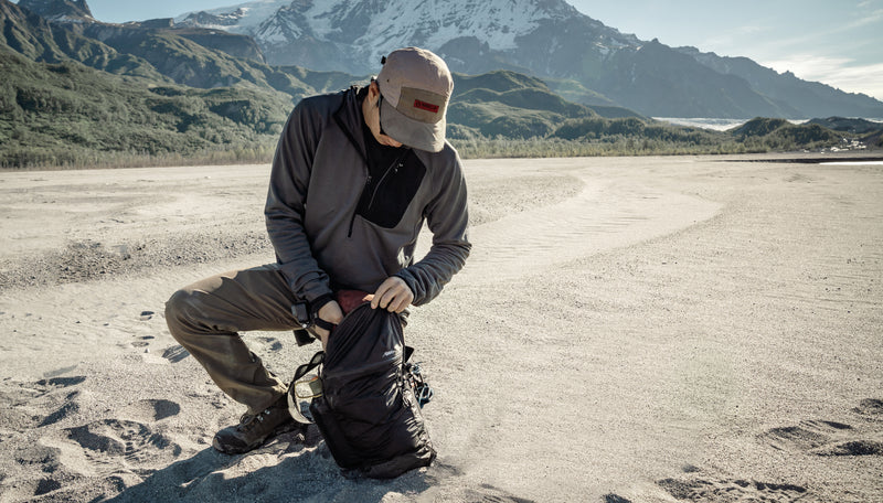 Man kneeling on sandy Alaskan shore, digging into Freefly16 backpack