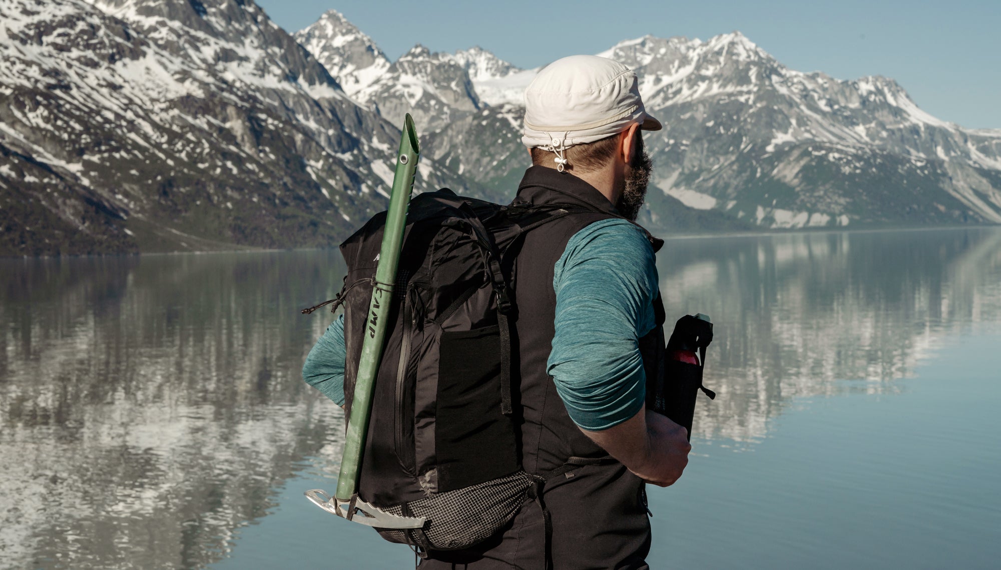 Back view of man wearing backpack in front of Alaskan lake with snow-covered peaks