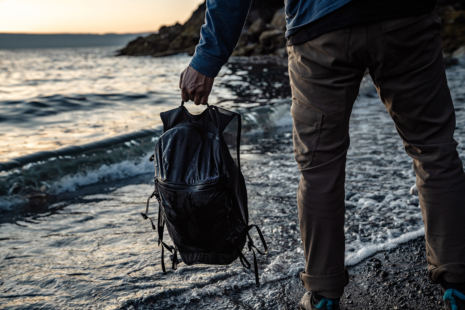 person holding the handle of a weatherproof travel backpack at a black sand beach