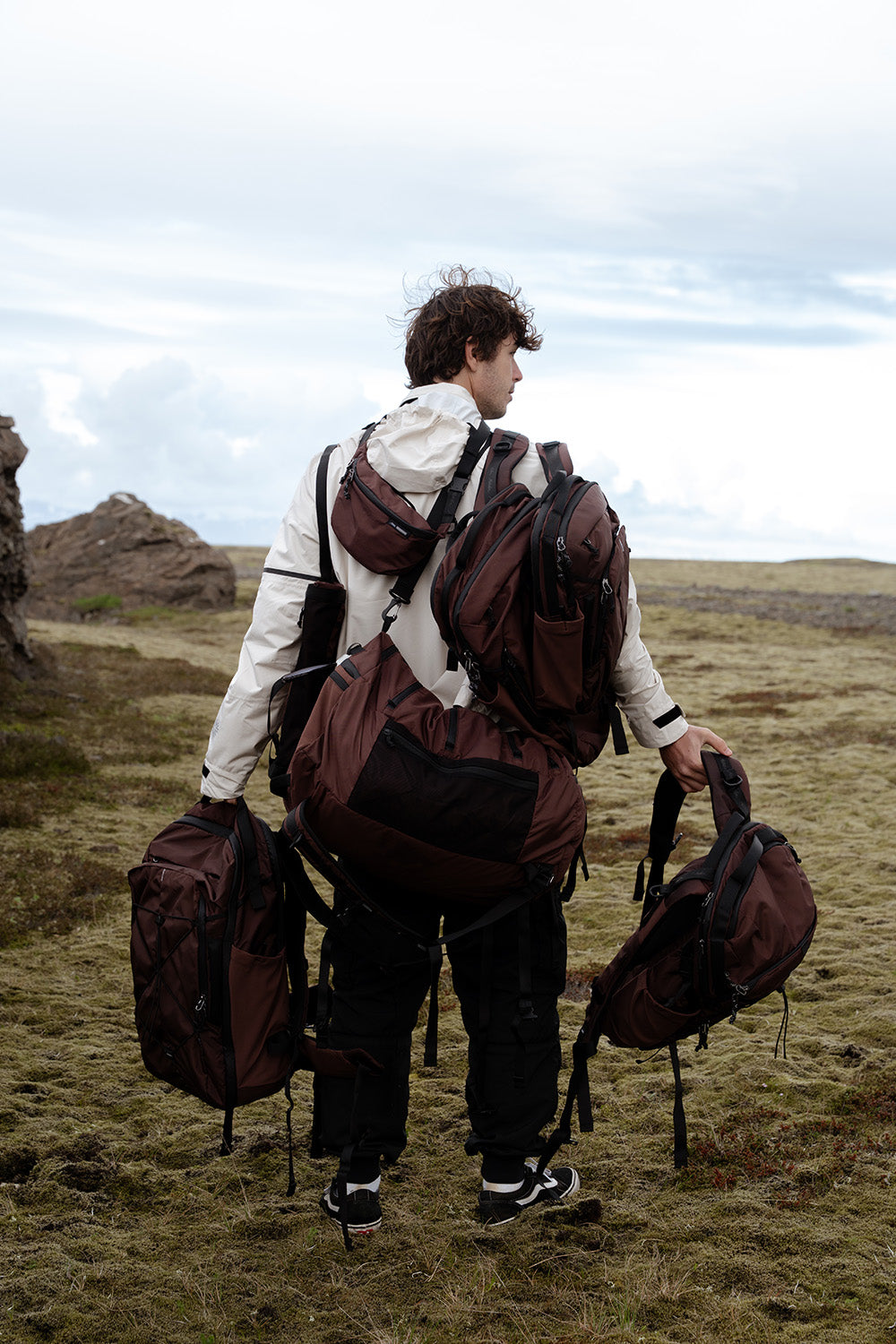 A person from behind in an open grassy landscape, wearing several backpacks, bags, and slings in matching garnet color.