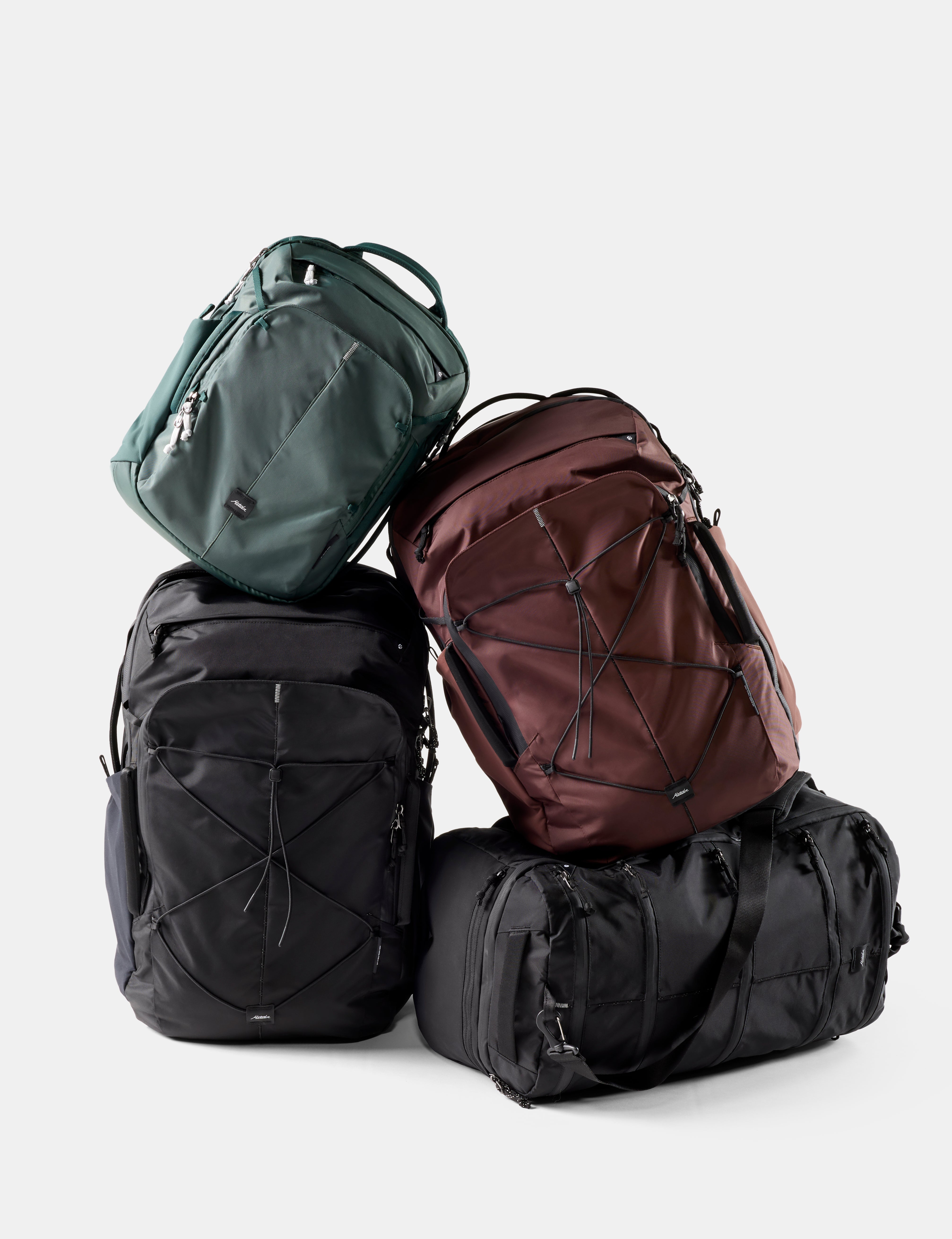 Three backpacks and a duffel bag arranged in a stacked pile on a light gray studio background. The set includes a dark green backpack on top, a black backpack underneath it, a dark red backpack on the right, and a black duffel bag at the bottom.