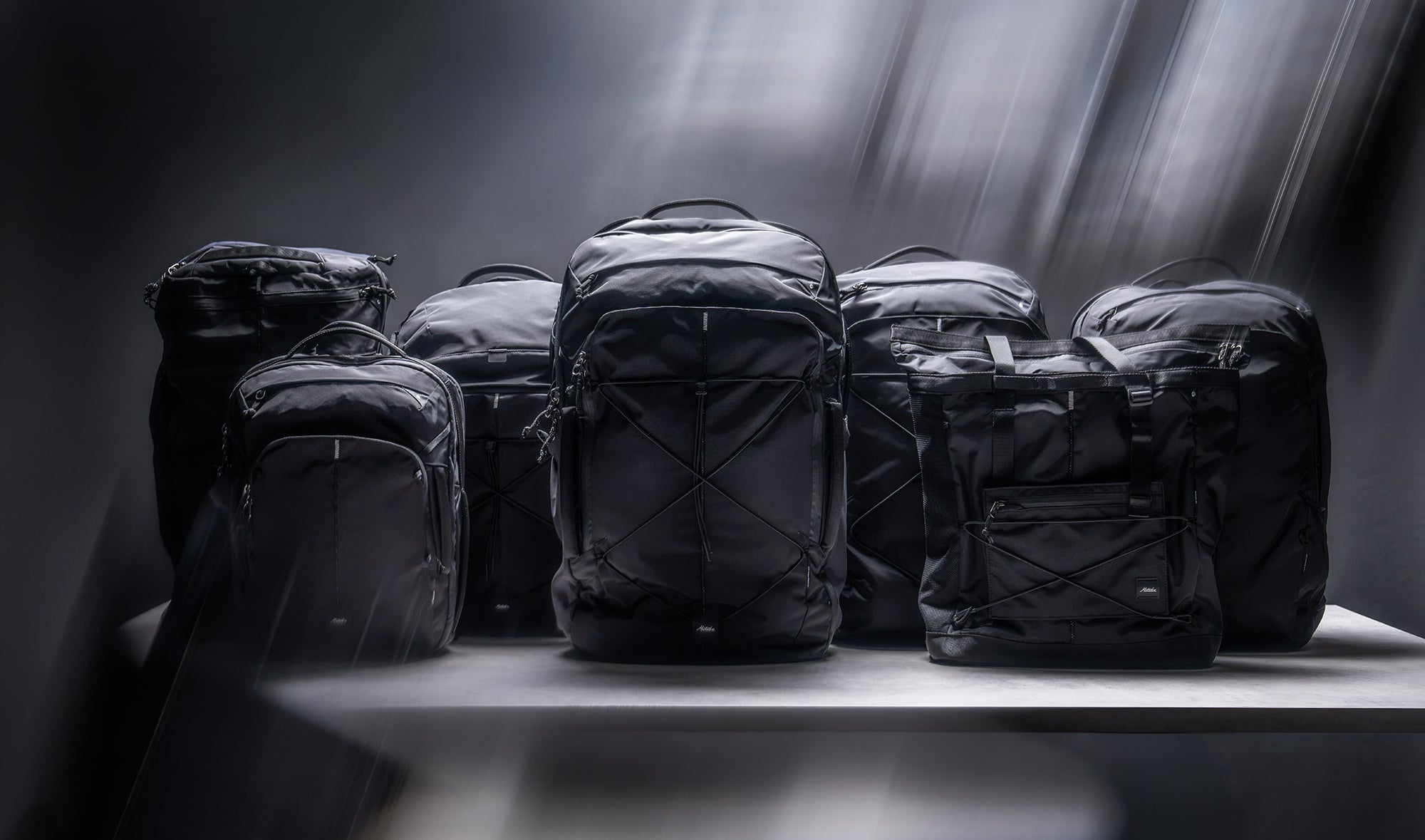 A lineup of black travel carry-on backpacks and bags displayed on a white table against a dark, moody studio background with light rays shining down on the bags.