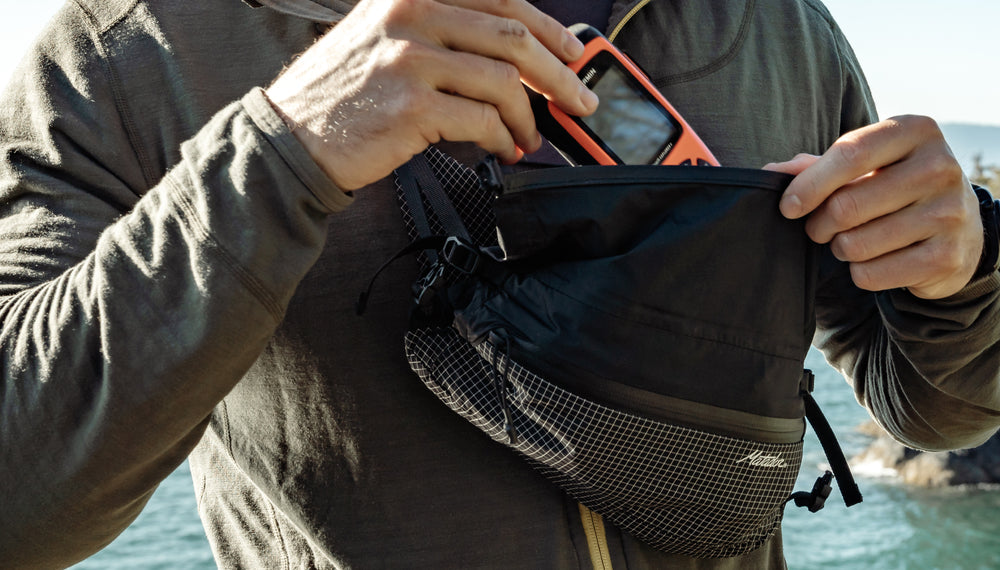 Man standing at shoreline, wearing cross body hip pack with orange GPS device