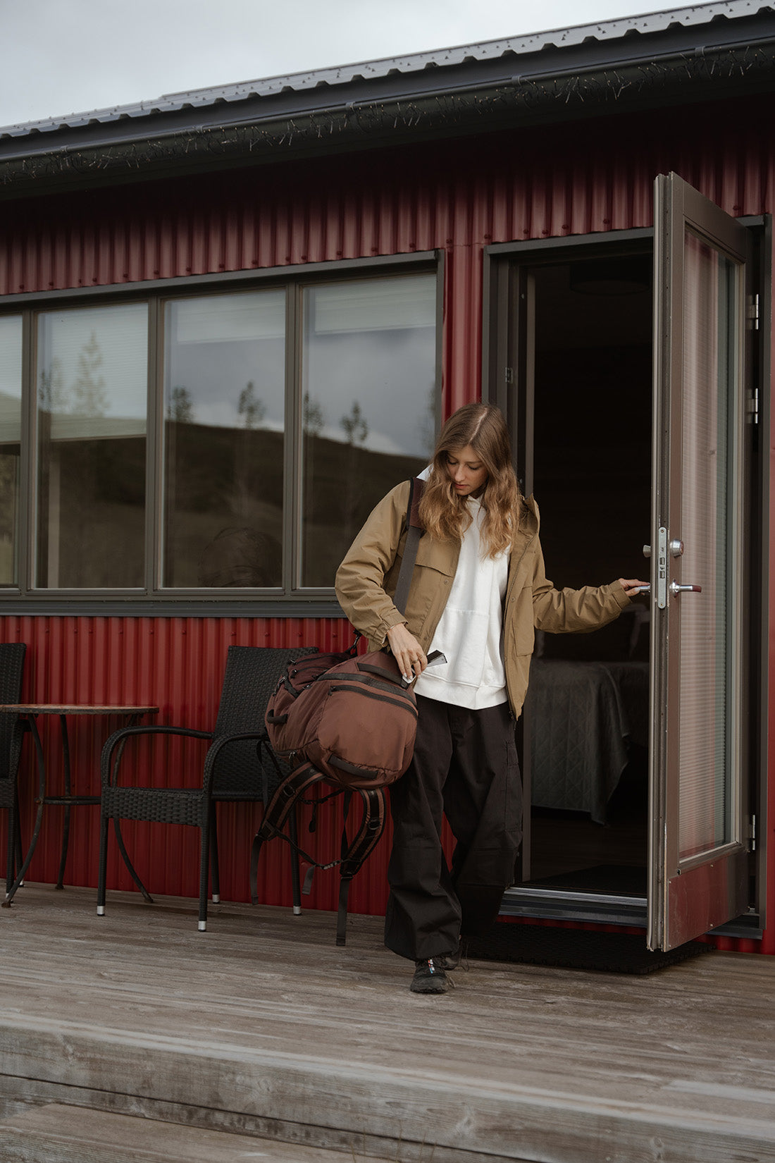 A woman stands at the open doorway of a red cabin, holding a garnet duffel bag and wearing a tan jacket, white shirt, and dark pants.