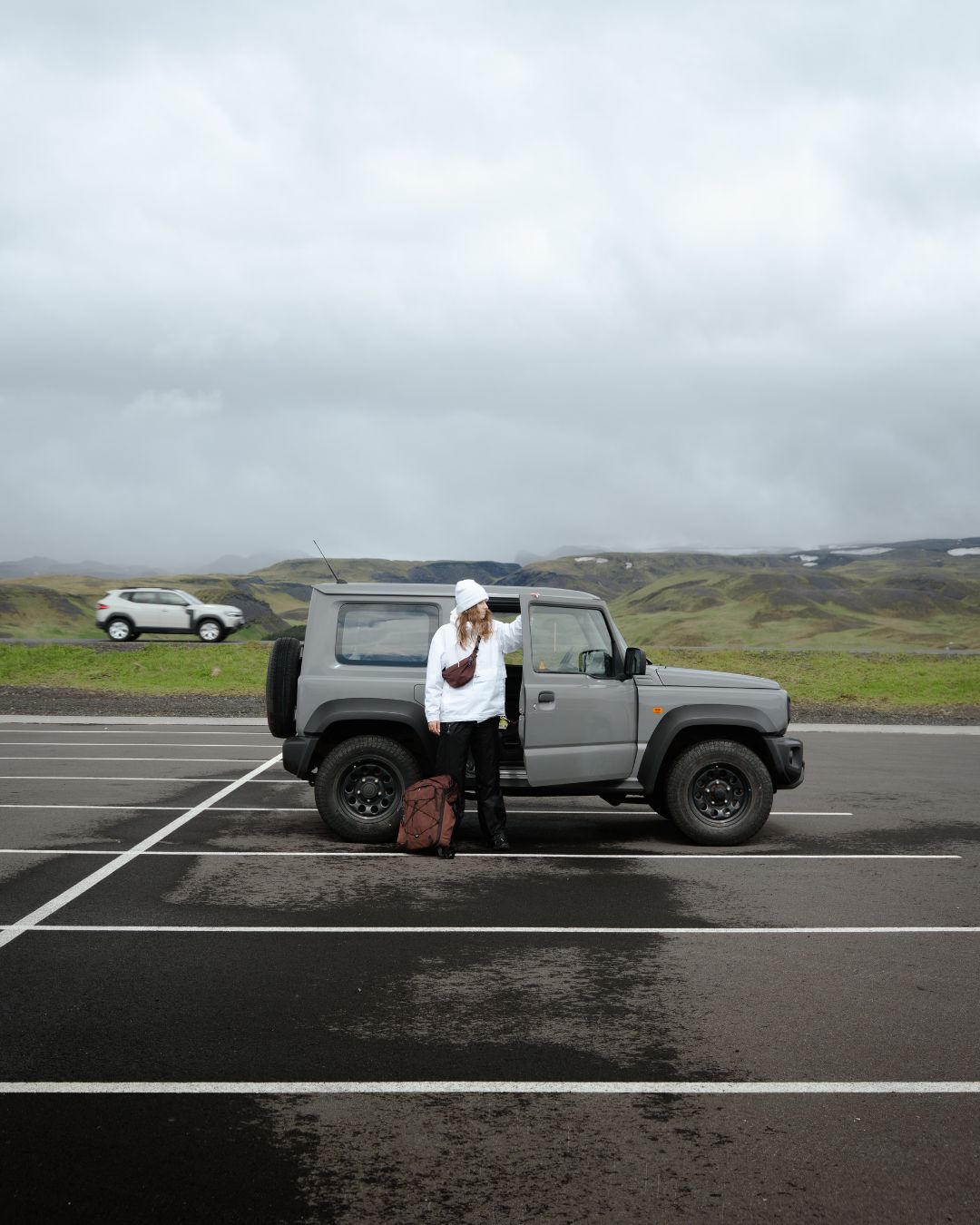 A woman in a white hoodie with a red backpack and cross-body sling bag and black pants stands next to a gray SUV parked in an outdoor lot under an overcast sky.
