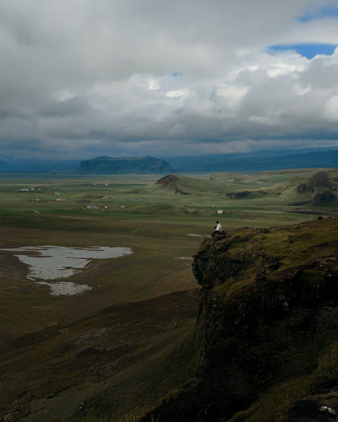 A dramatic landscape photograph of a vast green valley and distant mountains under a heavy, cloudy sky, with a small figure standing on a dark rocky cliff in the foreground.