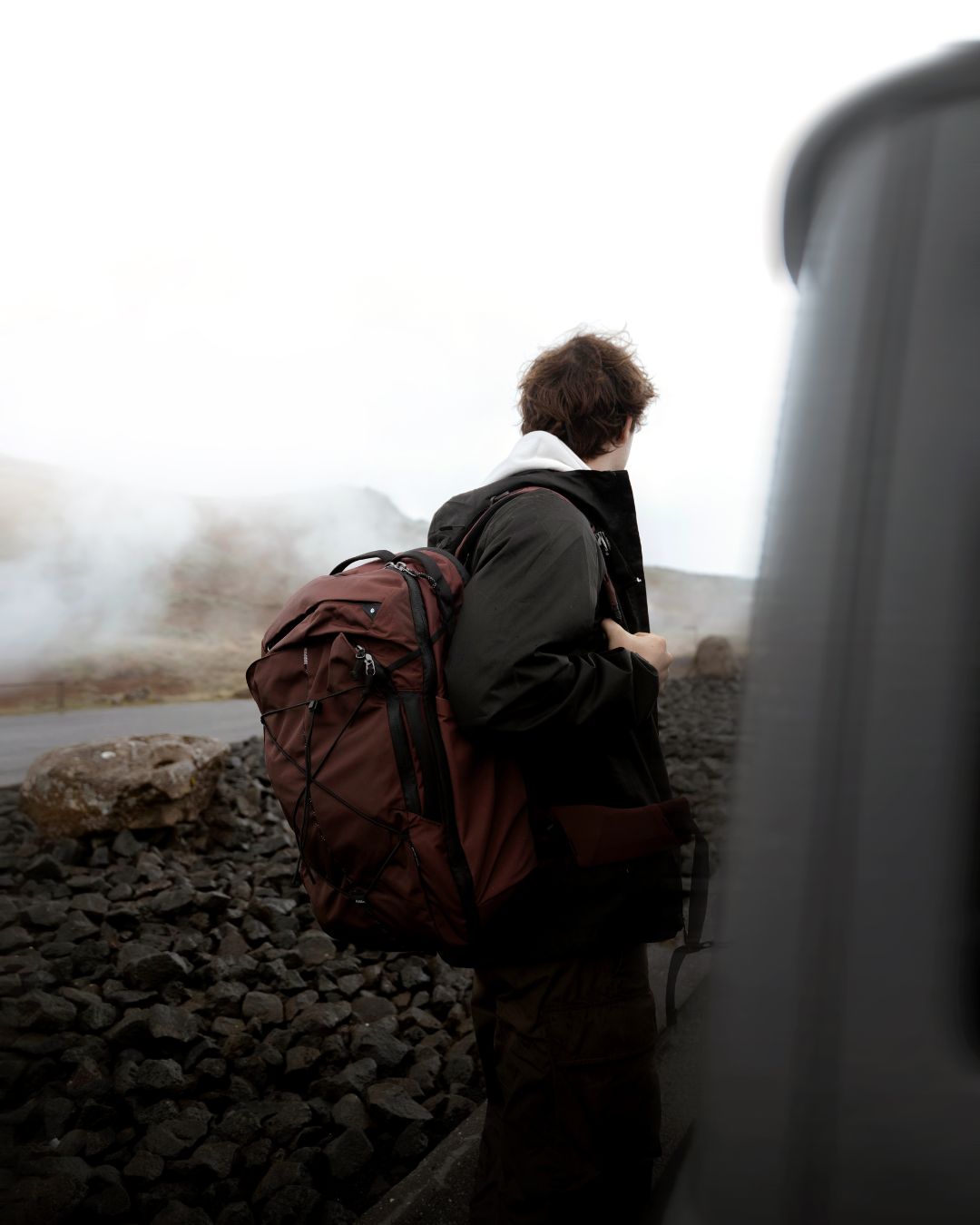 A person seen from behind wearing a dark jacket and carrying a large red hiking backpack, standing outdoors near a vehicle with rocks and mist in the background.