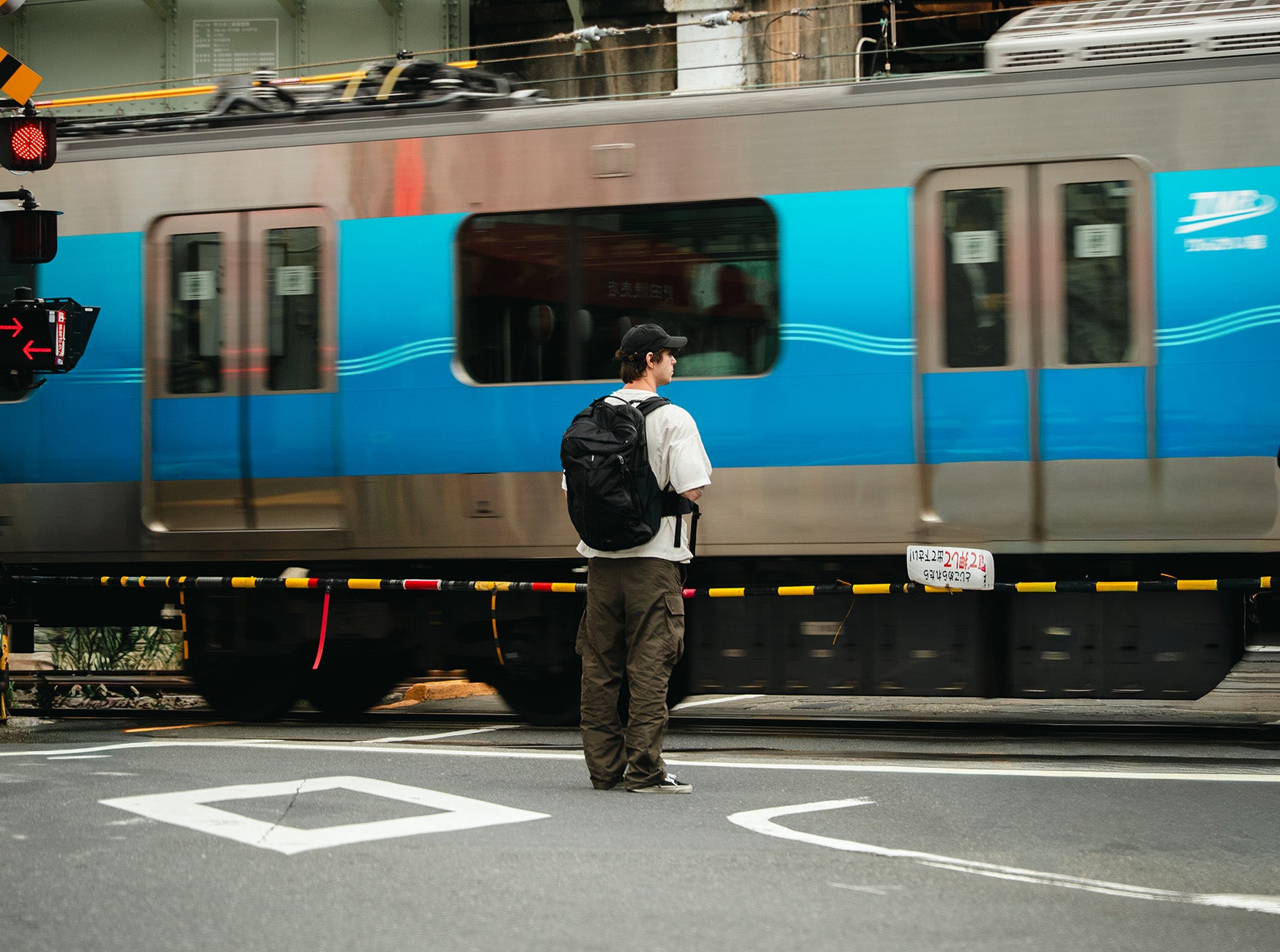 A man with a black travel bag wearing a white t-shirt, brown cargo pants, and a black backpack stands at a train station crossing, looking at a blue and silver train passing by.