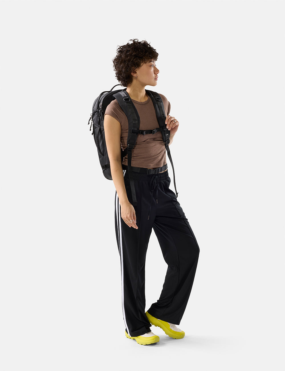 Person wearing a brown shirt, black pants with white stripes, and yellow shoes, holding a black backpack on a white background.