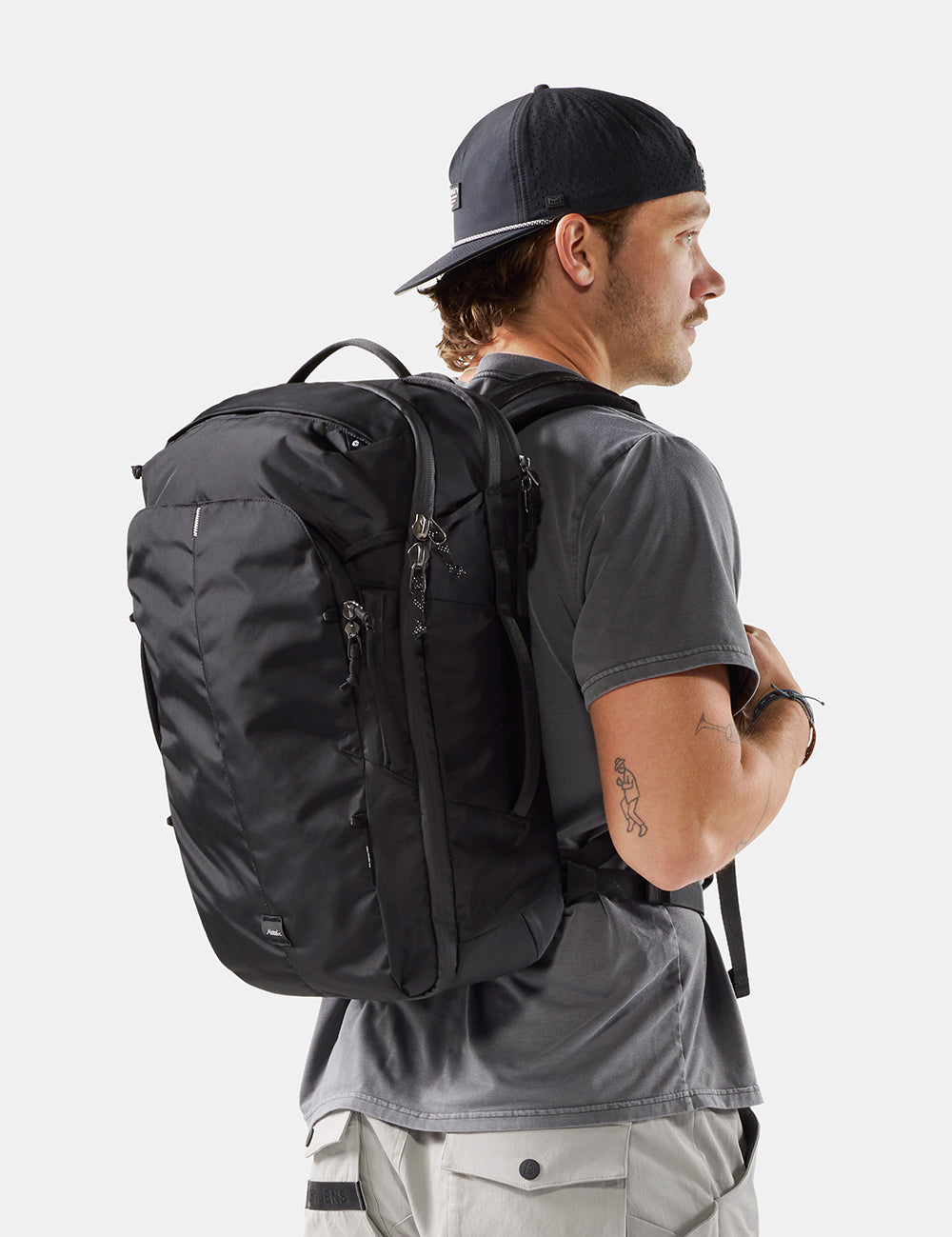 A man wearing a black baseball cap, gray t-shirt, and light gray cargo pants is shown from a side-back angle, wearing a black technical backpack. The photo is taken against a clean, light gray studio background.