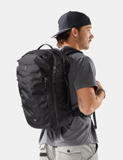 A man wearing a black baseball cap, gray t-shirt, and light gray cargo pants is shown from a side-back angle, wearing a black technical backpack. The photo is taken against a clean, light gray studio background.