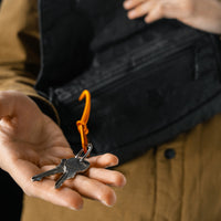 man holding keys out, connected to orange carabiner from black sling bag
