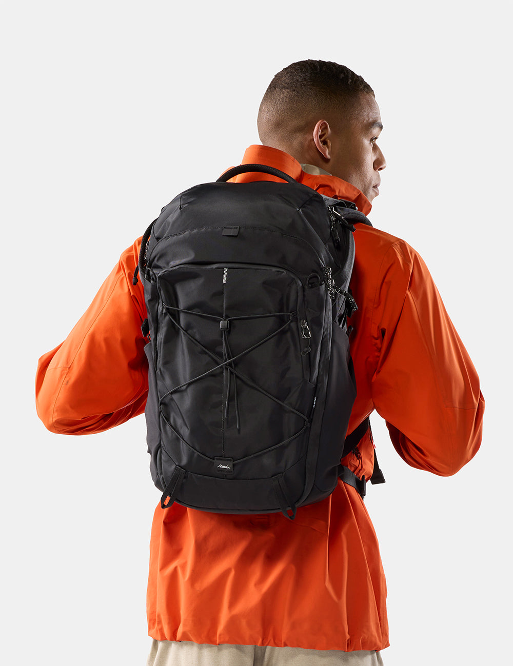 A man wearing a bright orange jacket is shown from the back, carrying a black technical backpack with crisscross compression straps on a light gray background.