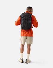 Person wearing an orange jacket and beige shorts with a black backpack on a white background
