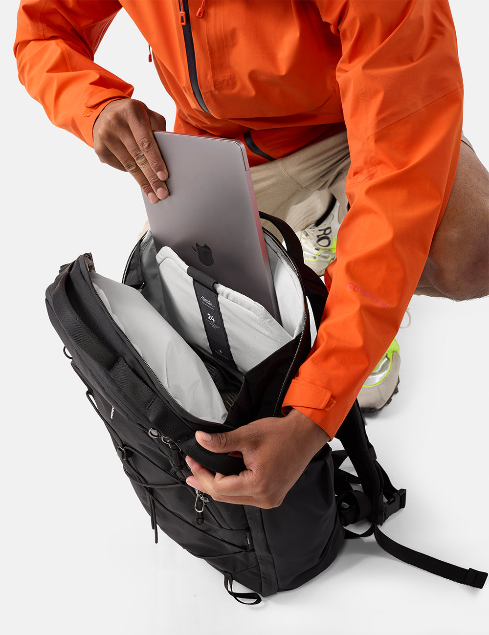 Person in orange jacket placing a laptop into a black backpack on a white background