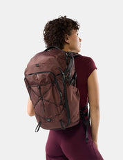 A woman wearing a garnet hiking backpack with black straps, shown from a rear three-quarter view against a light gray studio background.