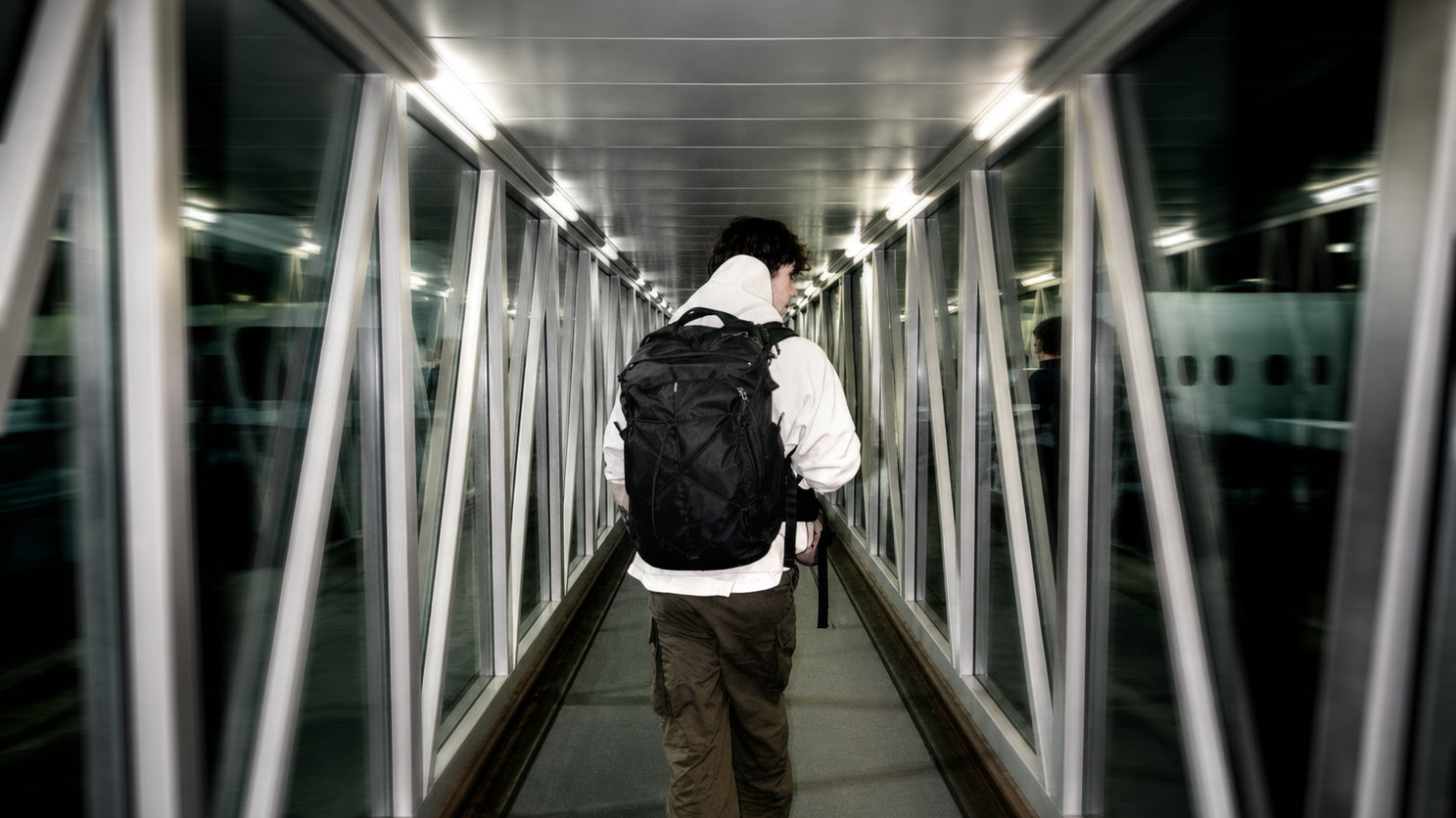 A person seen from behind walking through a glass-paneled airport, wearing a white hoodie, brown pants, and a black carry-on backpack.
